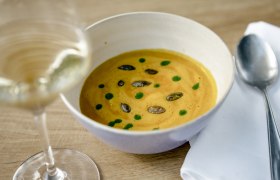 Eine Schüssel Kürbissuppe mit Kürbiskernen und grünen Tropfen, daneben ein Glas Weißwein und ein Löffel auf einer Serviette.