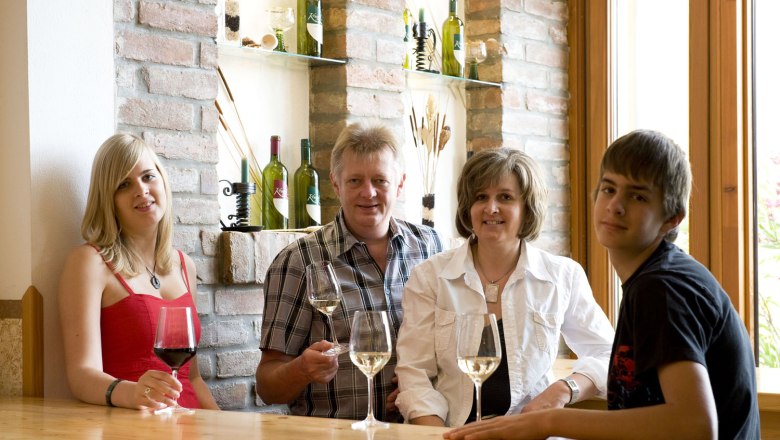 Vier Personen sitzen an einem Tisch mit Weingläsern, im Hintergrund Weinflaschen auf Regalen.