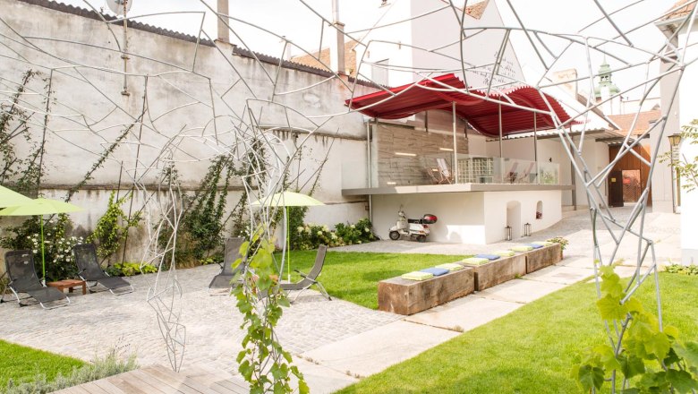 Moderner Innenhof mit Rasen, Liegest&uuml;hlen, Metallskulpturen und einer Vespa unter einem roten Vordach.