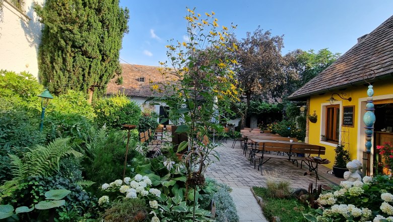 Ein gemütlicher Garten mit gelbem Gebäude, Holzbänken und üppiger Vegetation unter blauem Himmel.