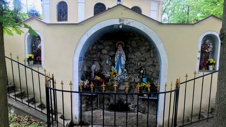 Eine Grotte mit religiösen Statuen und Blumen, umgeben von einem schmiedeeisernen Zaun.