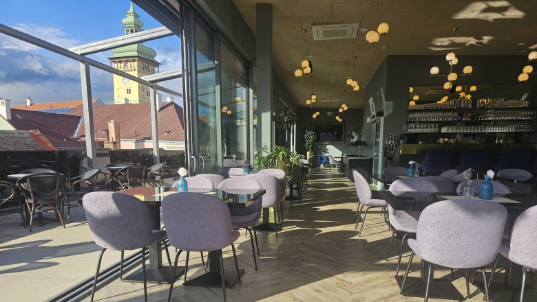 Innenbereich eines modernen Cafés mit Glaswänden, Blick auf den Rathausturm, Tische und Stühle, hölzerner Boden und dekorative Beleuchtung.