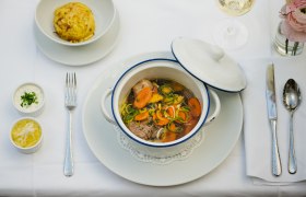 Ein gedeckter Tisch mit einem Eintopf in einem weißen Topf, einem Knödel, zwei Saucen und einem Glas Weißwein.