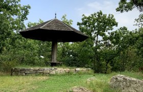 Ein hölzerner Pavillon mit spitzem Dach in einer grünen, bewaldeten Umgebung.
