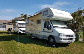 Ein weißes Wohnmobil mit ausgefahrener Markise steht auf einer Wiese neben einem weiteren Fahrzeug.