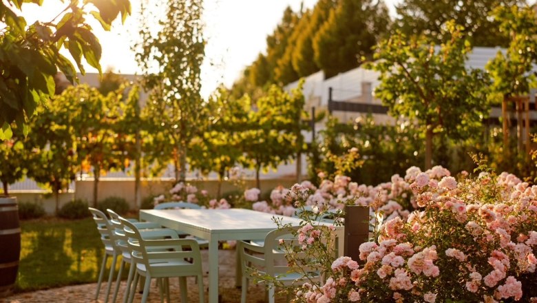 Ein Garten mit einem Tisch und Stühlen, umgeben von blühenden Rosen und Bäumen im Sonnenlicht.