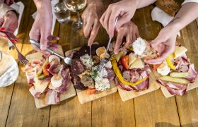 Mehrere Hände greifen nach Wurst- und Käseplatten auf einem Holztisch.