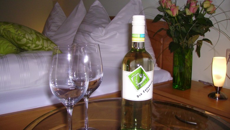 Ein gem&uuml;tliches G&auml;stezimmer mit einer Flasche Wein, zwei Gl&auml;sern und einem Blumenstrau&szlig; auf einem Tisch.