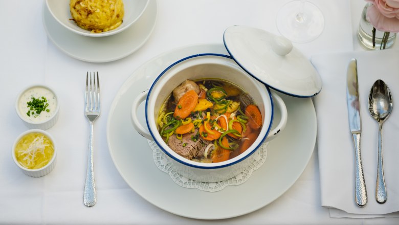 Ein gedeckter Tisch mit einem Eintopf in einem weißen Topf, einem Knödel, zwei Saucen und einem Glas Weißwein.