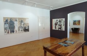 Innenansicht einer Kunstgalerie mit Holzboden, Gem&auml;lden an den W&auml;nden und einem Holztisch in der Mitte.