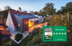 Ein modernes Kellerstöckl im Weinviertel bei Dämmerung, umgeben von Weinreben und einem Garten. Ein Schild zeigt 'Urlaub am Bauernhof'.