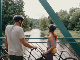 Zwei Radfahrer mit Helmen stehen auf einer Br&uuml;cke und blicken auf einen Fluss, umgeben von &uuml;ppiger Vegetation.