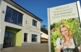 Ein modernes Gebäude mit einem Schild, das eine Frau in Tracht mit einem Weinglas zeigt. Text: Herzlich willkommen bei der Weinkönigin Tanja Dworzak.