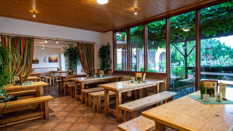 Innenansicht eines Heurigenlokals mit Holztischen und B&auml;nken, bunten Vorh&auml;ngen und gro&szlig;en Fenstern mit Blick auf einen begr&uuml;nten Au&szlig;enbereich.