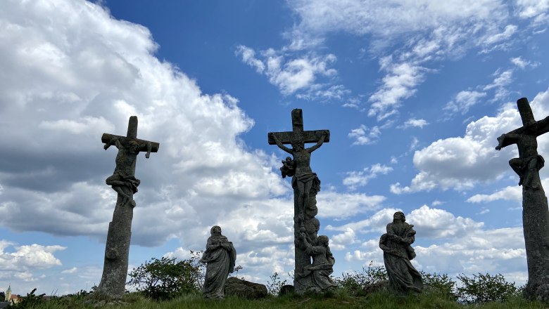 Drei steinerne Kreuze mit Figuren auf einem Hügel unter bewölktem Himmel.