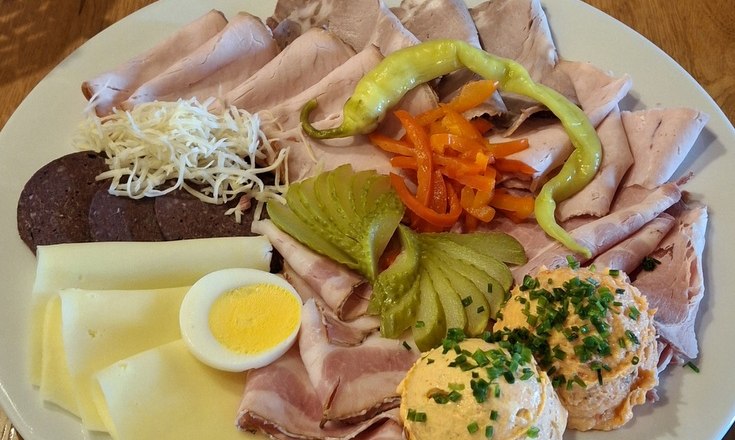 Ein Jausenteller mit verschiedenen Wurst- und Käsesorten, Ei, Paprika, Gurken und Aufstrich auf einem Holztisch.