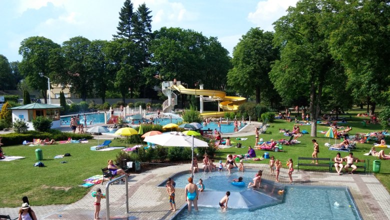 Ein Freibad mit mehreren Pools, einer gelben Wasserrutsche und vielen Menschen, die auf der Wiese entspannen.