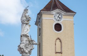 Kirchturm mit Uhr und Mariens&auml;ule in Schrattenthal vor blauem Himmel.