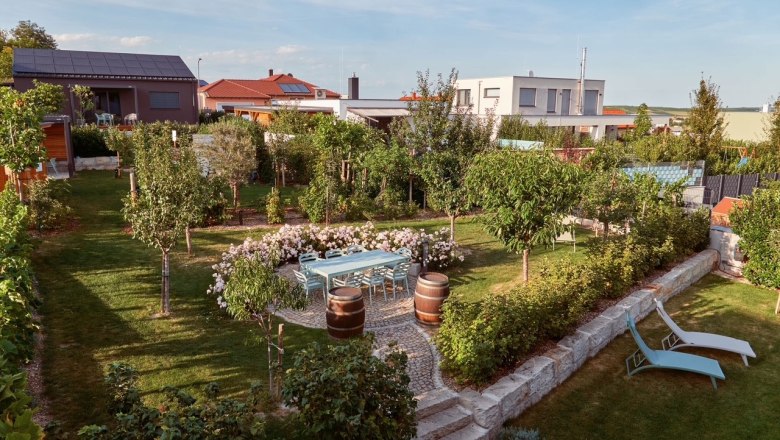 Ein gepflegter Garten mit Bäumen, Blumen, einem Tisch mit Stühlen und zwei Liegestühlen. Im Hintergrund sind moderne Häuser zu sehen.