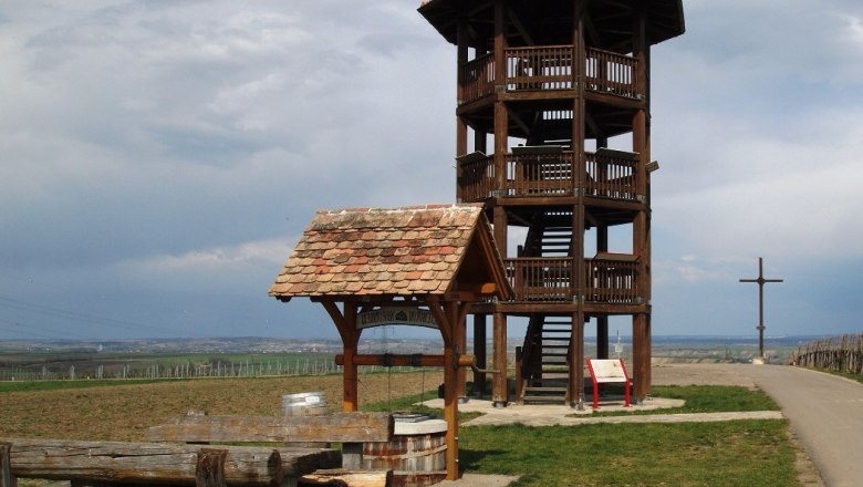 Aussichtsturm aus Holz auf einer Wiese mit Sitzbänken und einem Kreuz im Hintergrund.