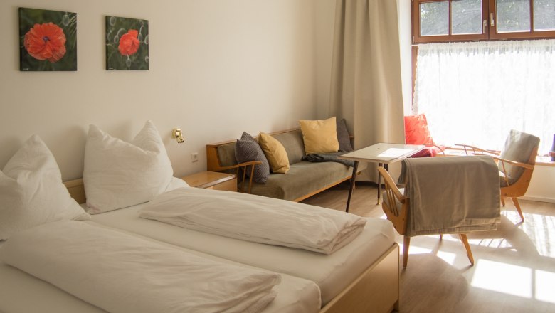 Ein gem&uuml;tliches Schlafzimmer mit Doppelbett, Sofa, Tisch und St&uuml;hlen. Helle Vorh&auml;nge und zwei Blumenbilder an der Wand.