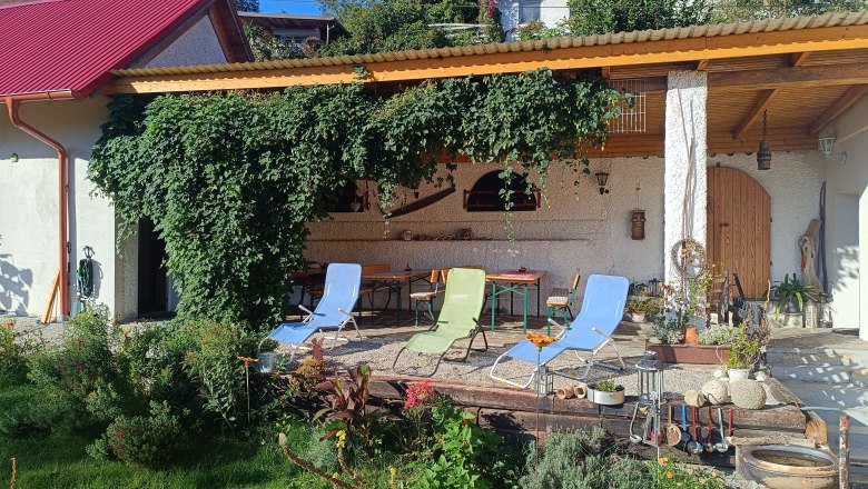 Gartenbereich mit Liegest&uuml;hlen und &uuml;berdachter Terrasse, umgeben von Pflanzen und Kletterpflanzen.