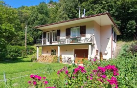 Ein wei&szlig;es Haus mit Balkon und Holzstapel im Garten, umgeben von gr&uuml;nen B&auml;umen und pinken Blumen im Vordergrund.