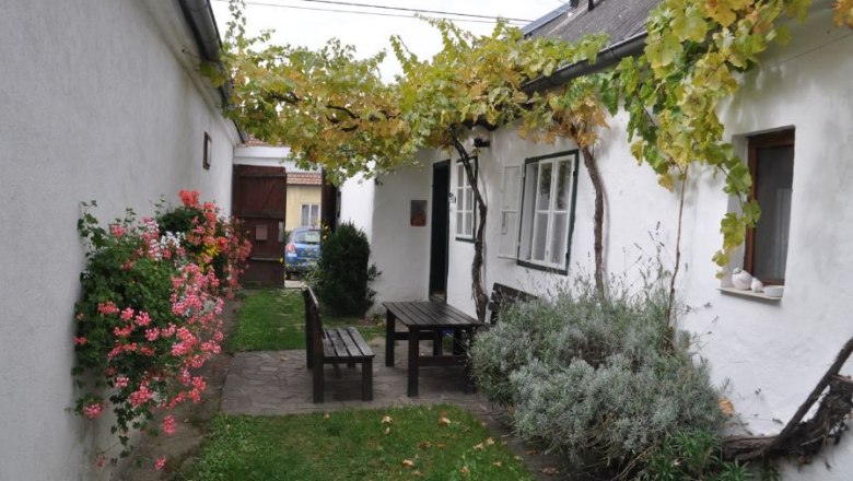 Innenhof, © Familie Ackerl Ein gemütlicher Innenhof mit Holztisch und Bänken, umgeben von weißen Wänden, Blumen und Weinreben.