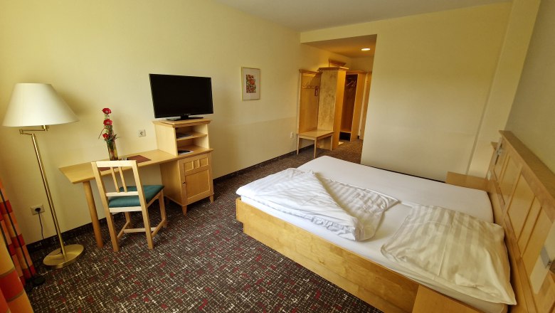 Ein einfach eingerichtetes G&auml;stezimmer mit Bett, Schreibtisch, Stuhl, Fernseher und Stehlampe.