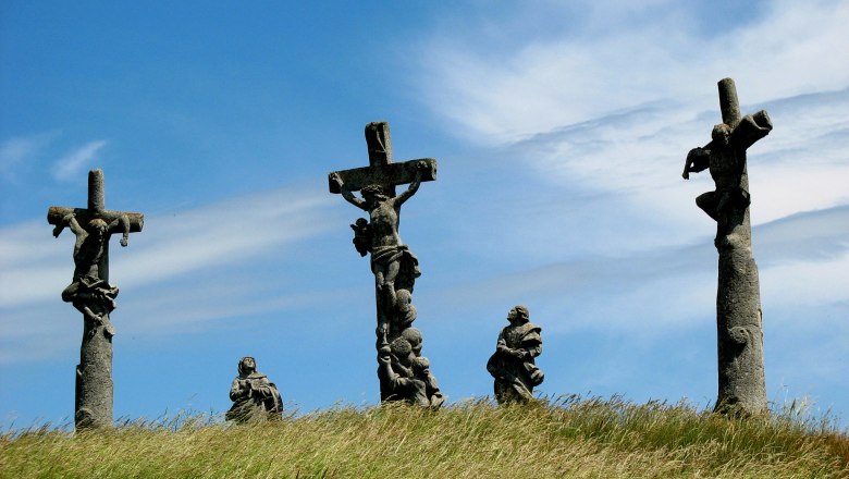 Drei Kreuze mit Figuren auf einem Hügel unter blauem Himmel.
