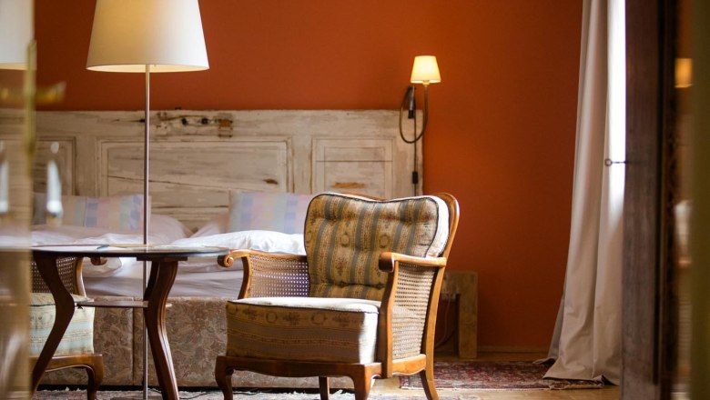 Gemütliches Zimmer mit Sessel, Tisch und Stehlampe vor einem Bett mit rustikaler Holzverkleidung.