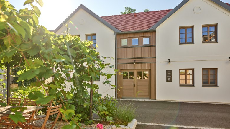 Ein modernes, zweistöckiges Gebäude mit weißen Wänden und roten Dachziegeln. Im Vordergrund ein Garten mit Holztisch, Stühlen und blühenden Pflanzen.