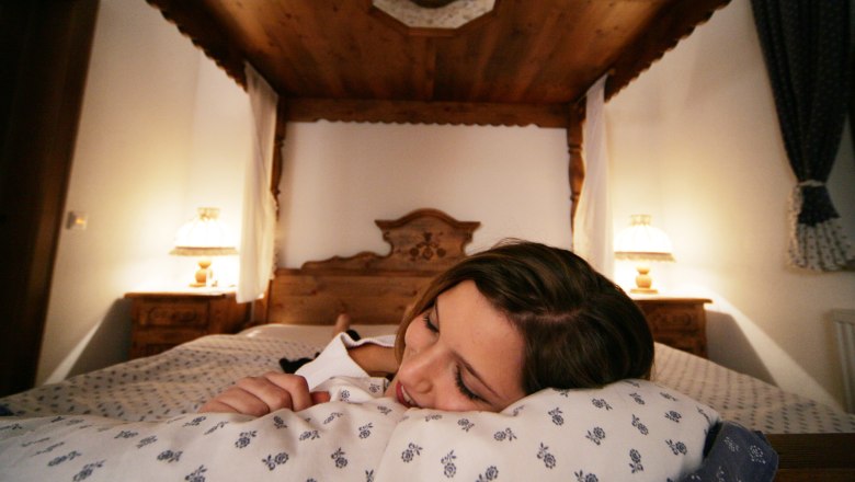 Herzlich Willkommen, © Weinviertel Tourismus GmbH / Weinfranz Frau liegt lächelnd auf einem Kissen in einem gemütlichen Hotelzimmer mit Holzdekor.