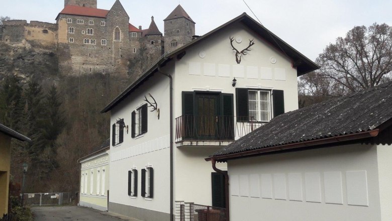 Ein wei&szlig;es Haus mit gr&uuml;nen Fensterl&auml;den und Hirschgeweihen an der Wand, im Hintergrund eine Burg auf einem H&uuml;gel.