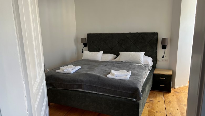 Schlafzimmer, © Balcar Ein gemütliches Schlafzimmer mit einem großen Bett, grauer Bettwäsche und weißen Handtüchern. Ein Kronleuchter hängt von der Decke.