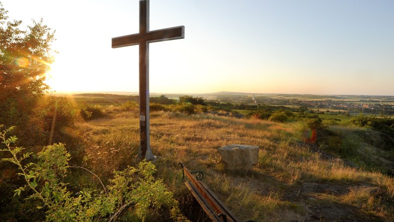 Ein Kreuz auf einem Hügel bei Sonnenuntergang mit Blick auf eine weite Landschaft und ein Dorf im Hintergrund.