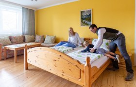 Ein gem&uuml;tliches Zimmer mit gelber Wand, einem Holzbett und einer Familie, die auf dem Bett spielt.
