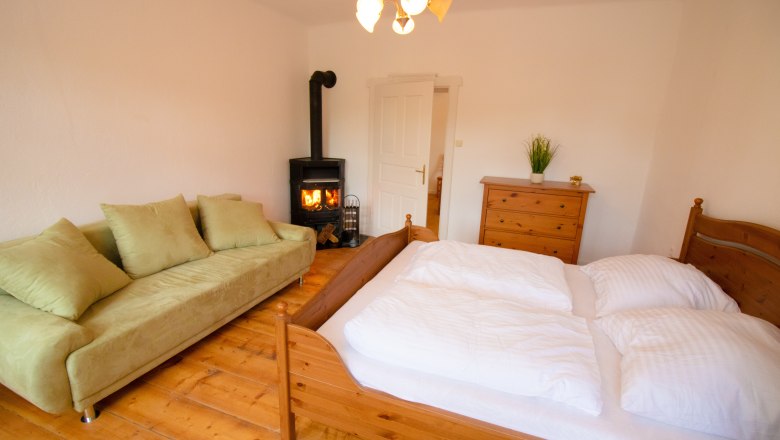 Gem&uuml;tliches Schlafzimmer mit Holzfu&szlig;boden, gr&uuml;nem Sofa, Holzofen, Doppelbett und Kommode.