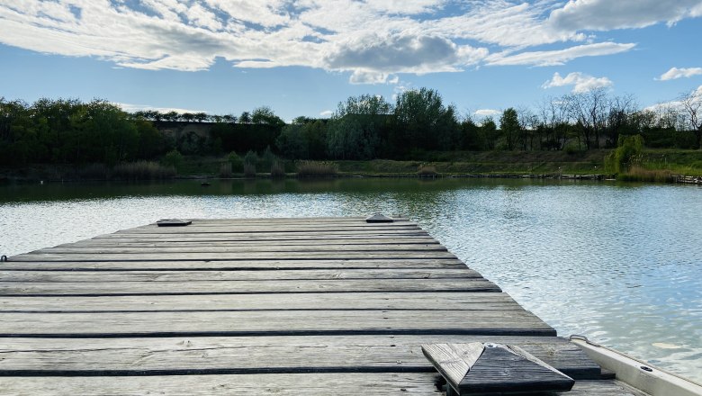 Holzsteg am Ziegelofenteich in Zellerndorf, umgeben von Bäumen und blauem Himmel mit Wolken.