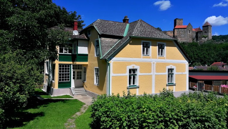 Gelbes Haus mit gr&uuml;nem Garten, im Hintergrund eine Burg auf einem H&uuml;gel.