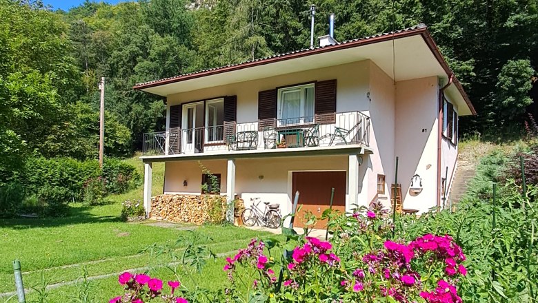 Ein wei&szlig;es Haus mit Balkon und Holzstapel im Garten, umgeben von gr&uuml;nen B&auml;umen und pinken Blumen im Vordergrund.