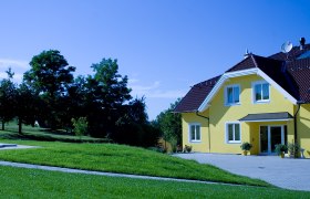 Gelbes Haus mit Garten und B&auml;umen im Hintergrund.
