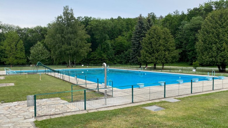 Ein leeres Freibad mit einem gro&szlig;en Schwimmbecken, umgeben von B&auml;umen und Rasenfl&auml;chen.