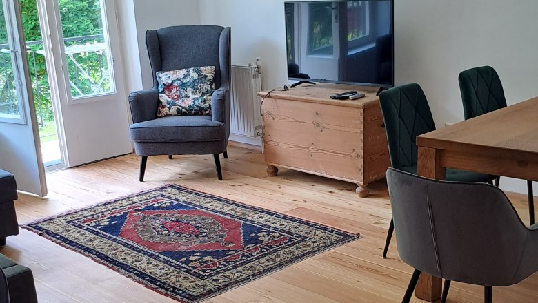 Wohnzimmer mit Holzboden, Teppichen, einem Sessel, einem Fernseher und einem Esstisch mit St&uuml;hlen.