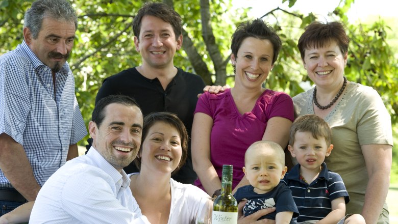 Eine Familie posiert lächelnd im Freien mit einer Flasche Wein auf einem Tisch.