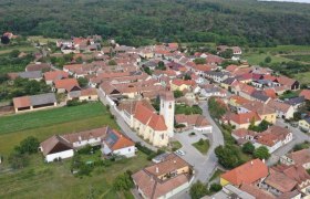Luftaufnahme eines kleinen Dorfes mit einer zentralen Kirche und roten D&auml;chern, umgeben von gr&uuml;nen Feldern und W&auml;ldern.
